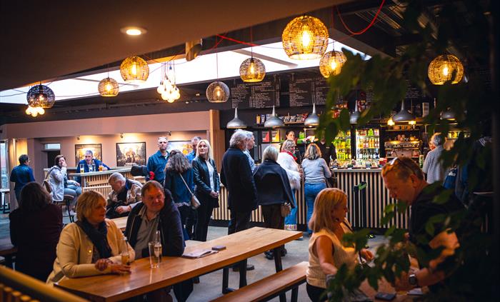 People sit and stand in a busy bar area with wooden tables and hanging lights